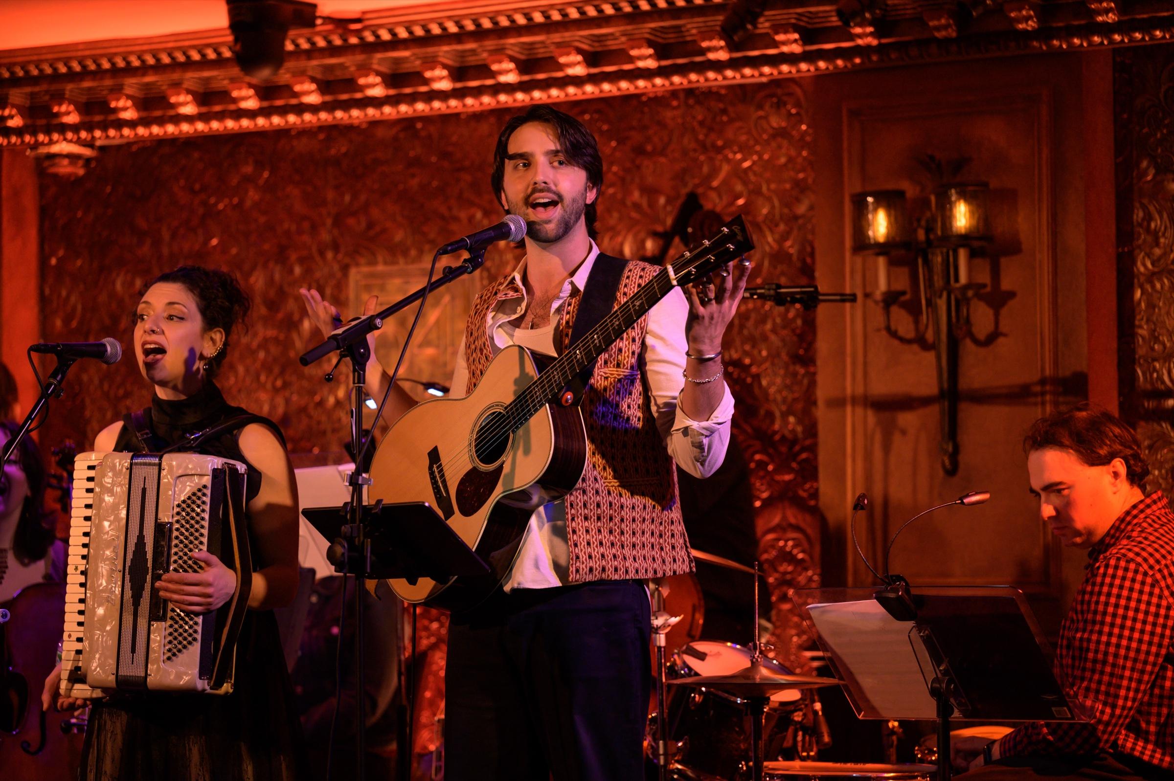 Shane Lonergan singing and playing acoustic guitar with the band at 54 Below, an accordionist beside him