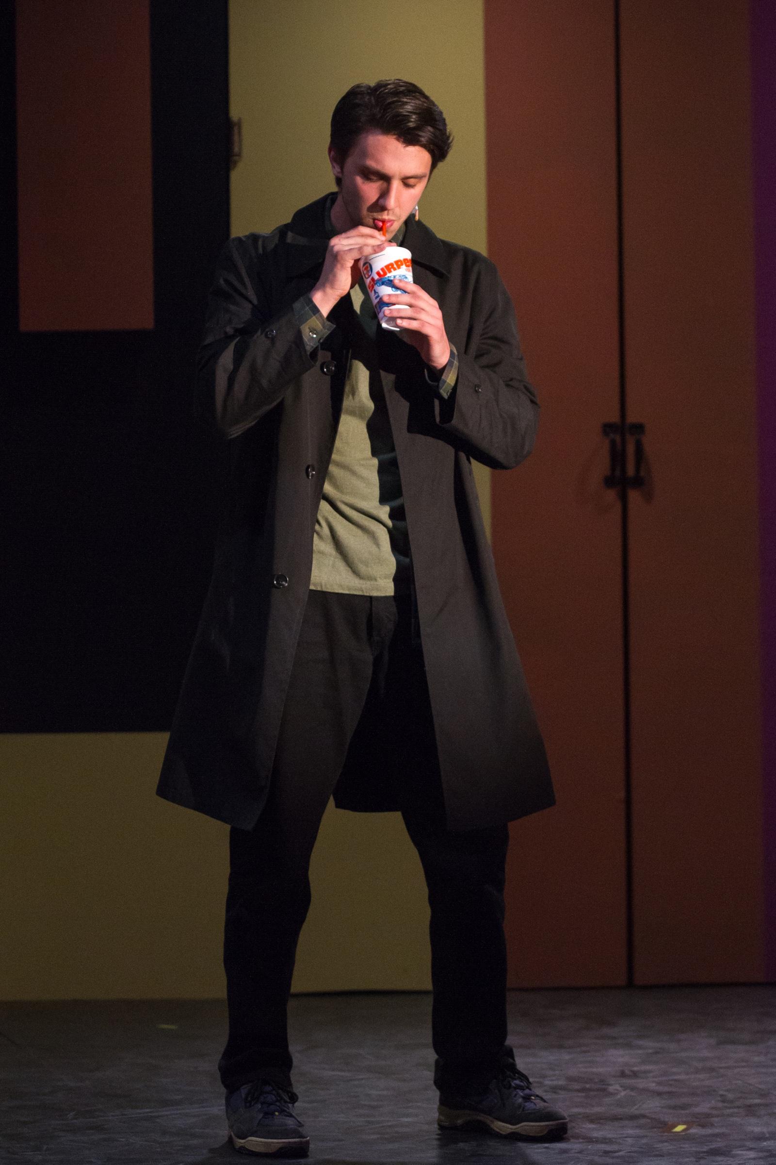 Shane Lonergan as J.D. in a long dark coat, drinking from a cup centre stage