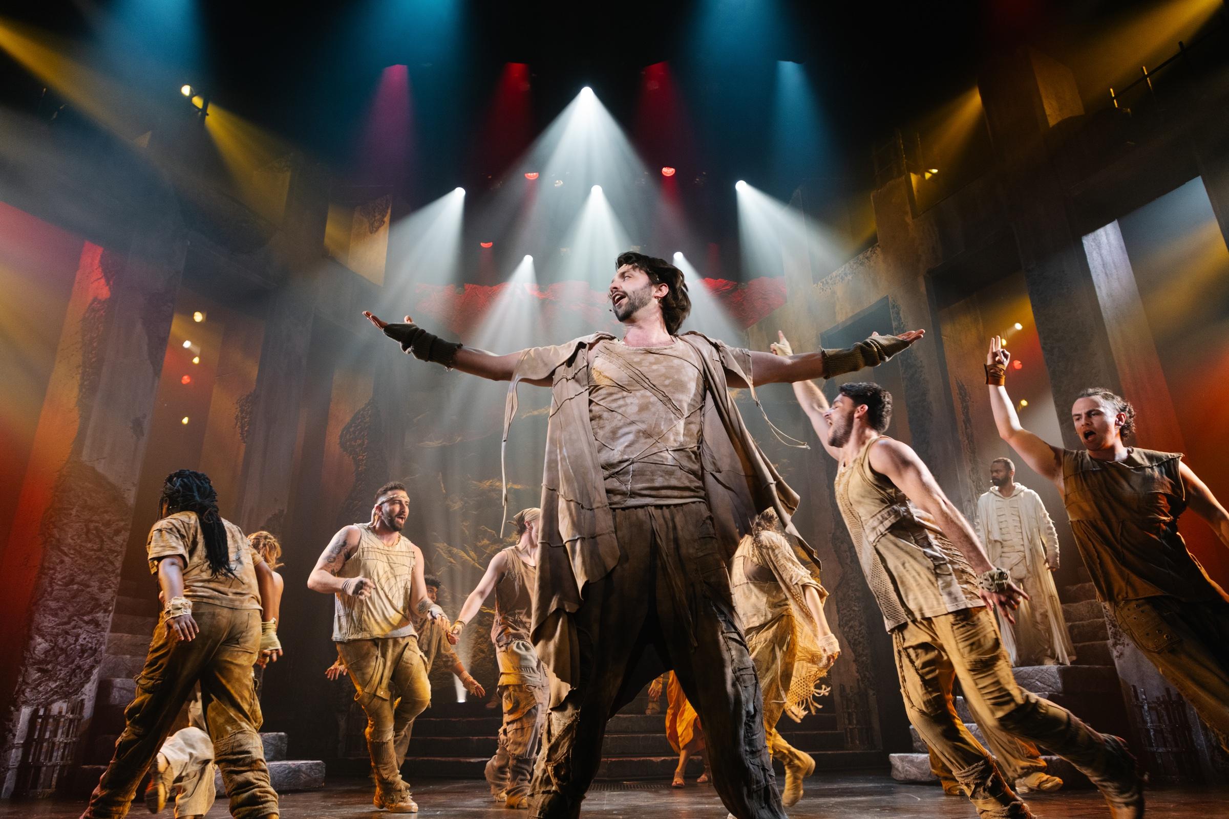Shane Lonergan centre stage, arms outstretched, with the company of Jesus Christ Superstar under a fan of coloured light