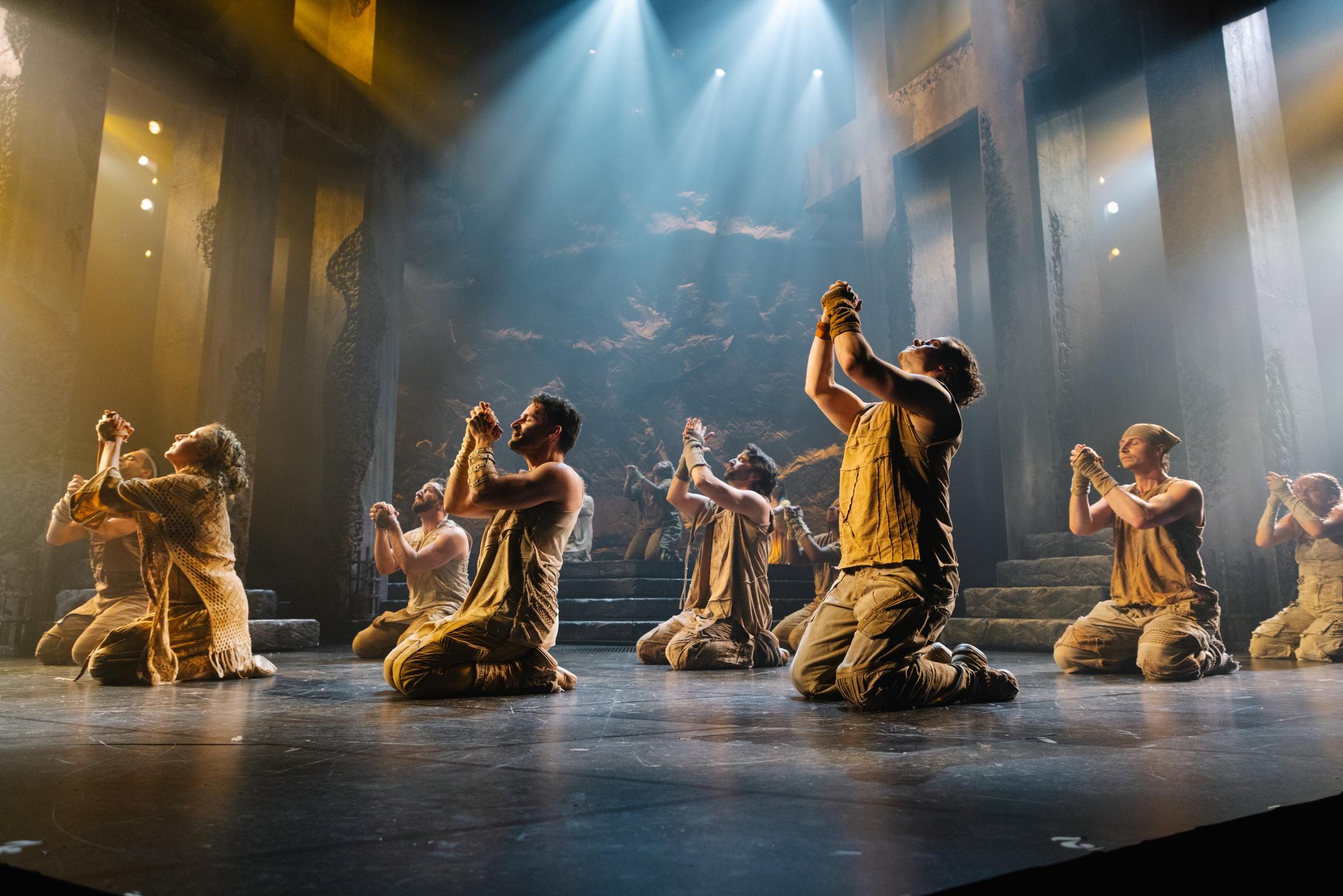 The company of Jesus Christ Superstar kneeling on a darkened stage as shafts of pale light fall from above