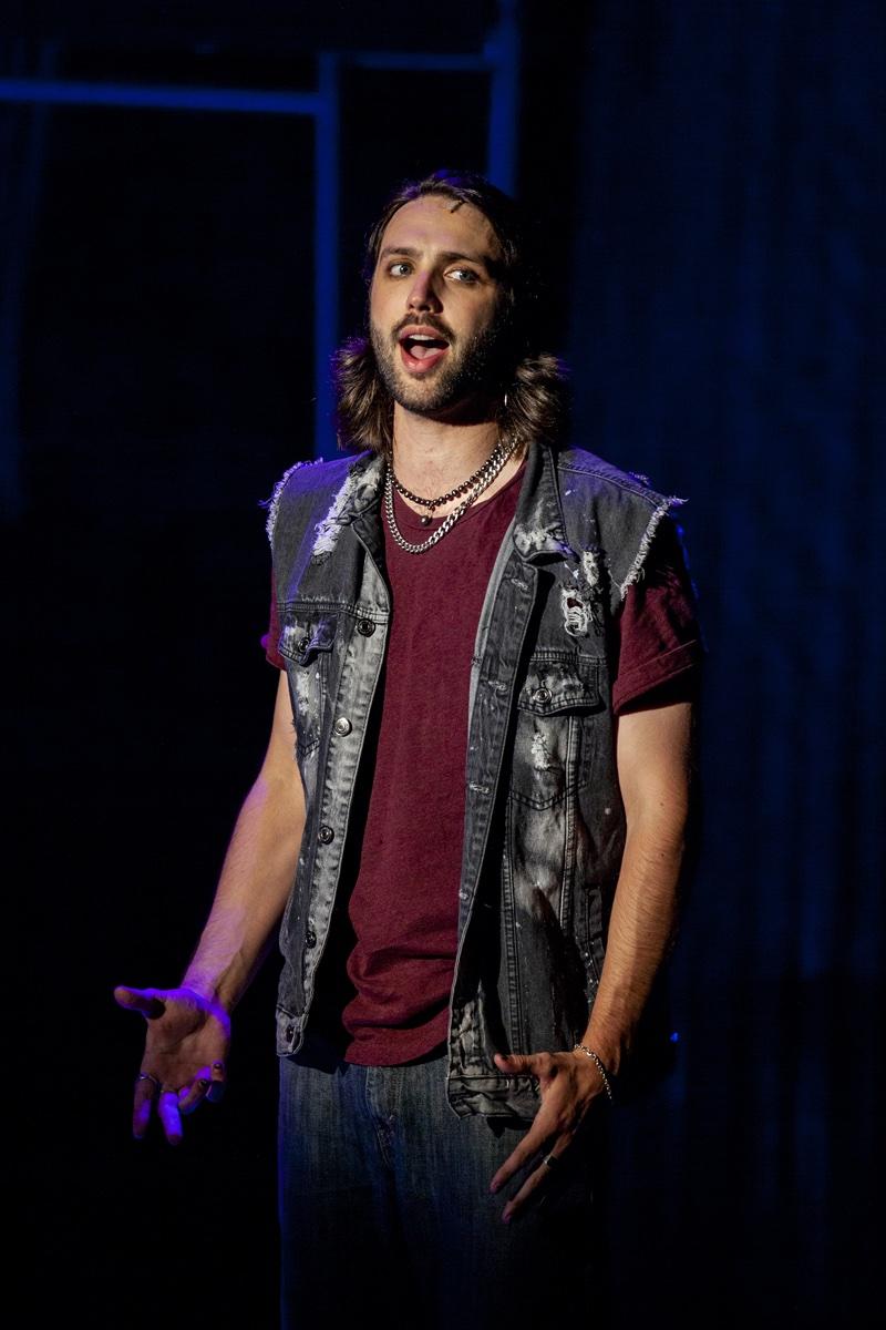 Shane Lonergan as Roger in a studded vest, singing under purple light