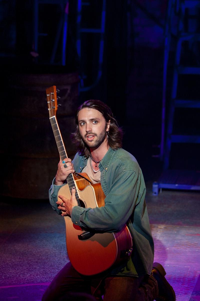 Shane Lonergan as Roger kneeling with an acoustic guitar in blue light
