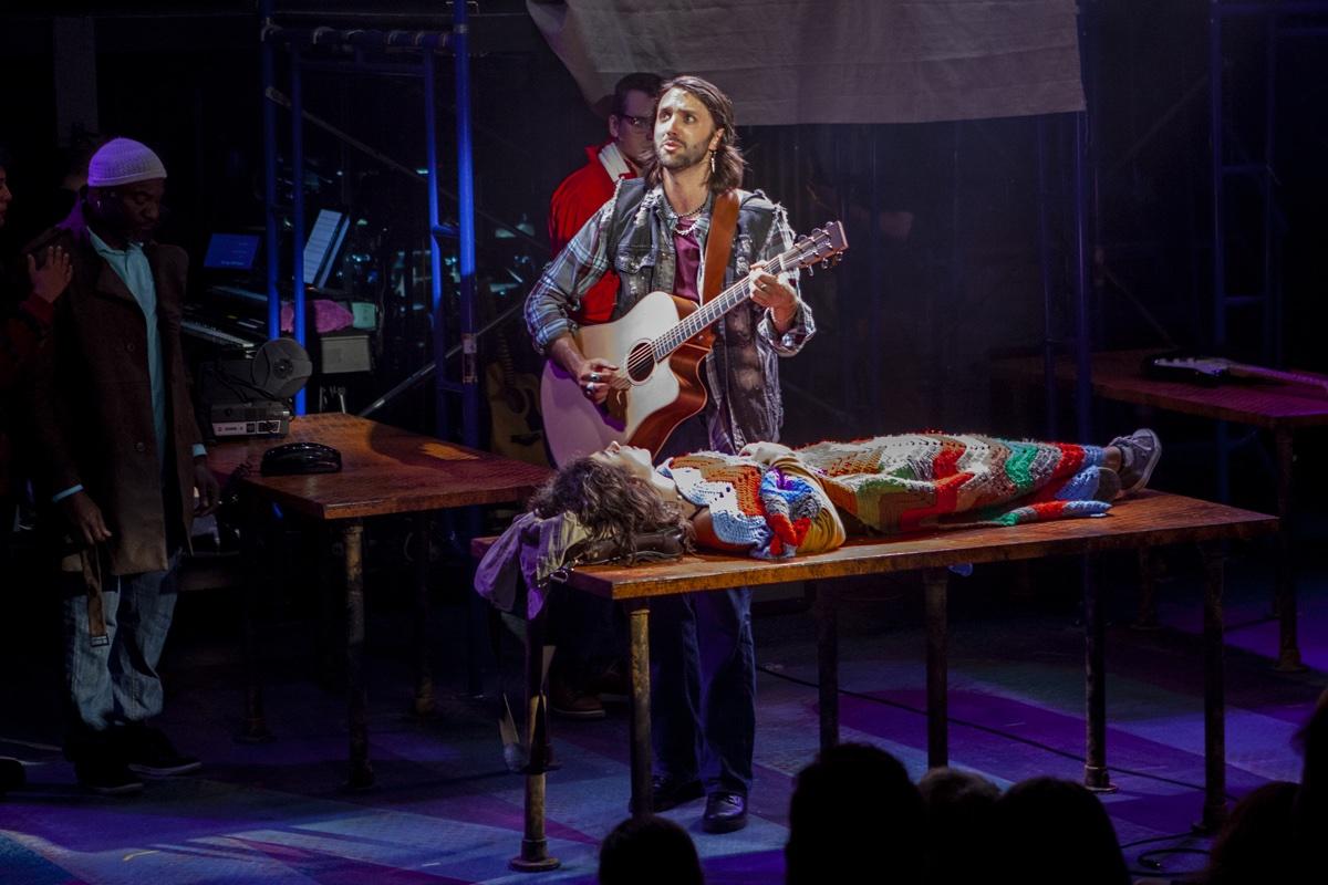 Shane Lonergan playing guitar beside a table where another performer lies in a patterned coat