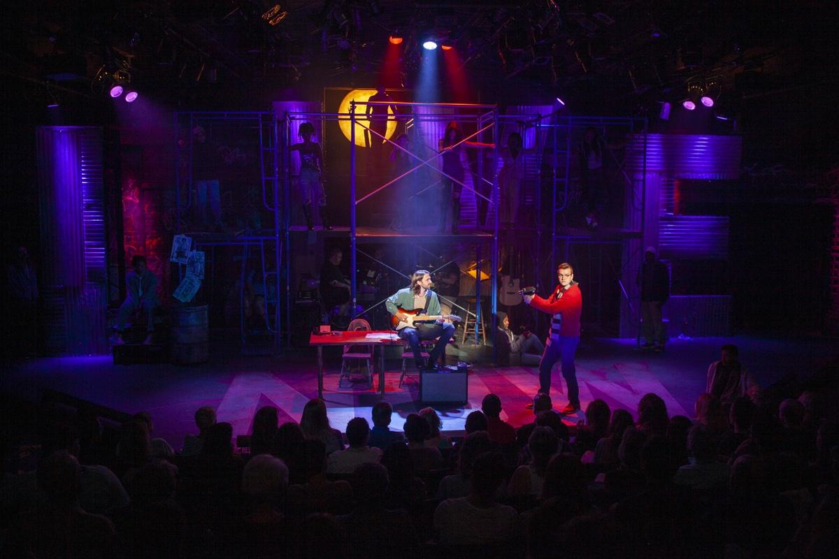 A wide view of the Rent stage in warm light, the silhouetted audience in the foreground