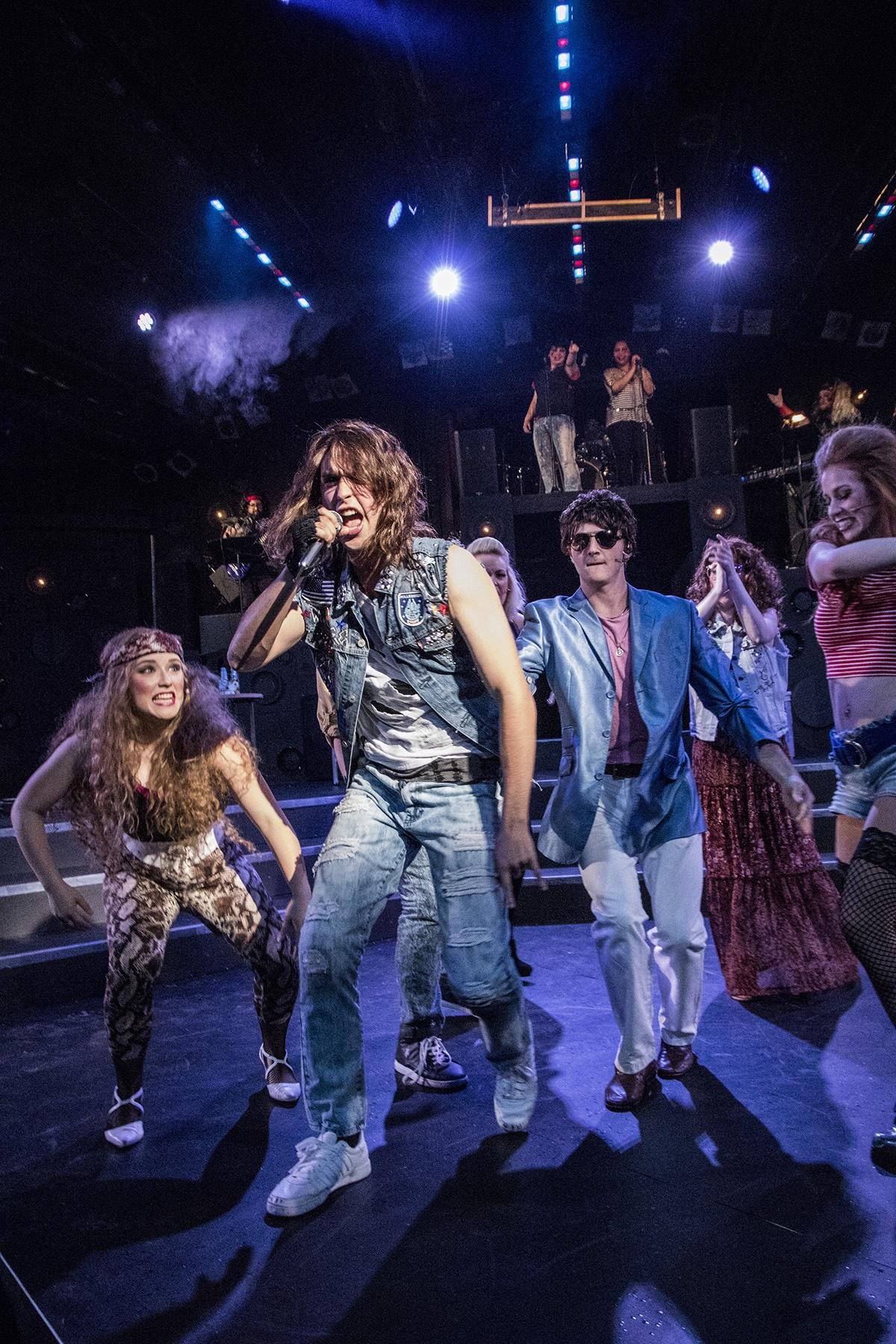 Shane Lonergan as Drew singing in denim under stage spotlights, the company behind him