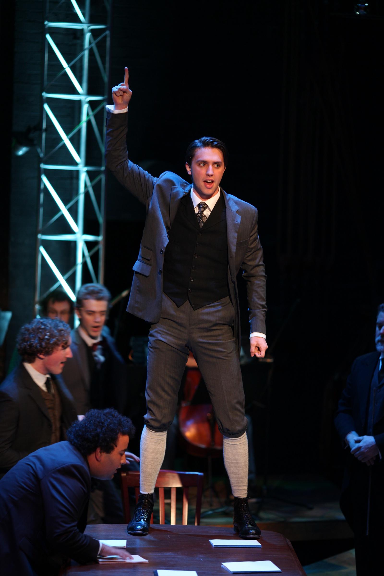 Shane Lonergan as Melchior standing above the seated boys, one arm raised
