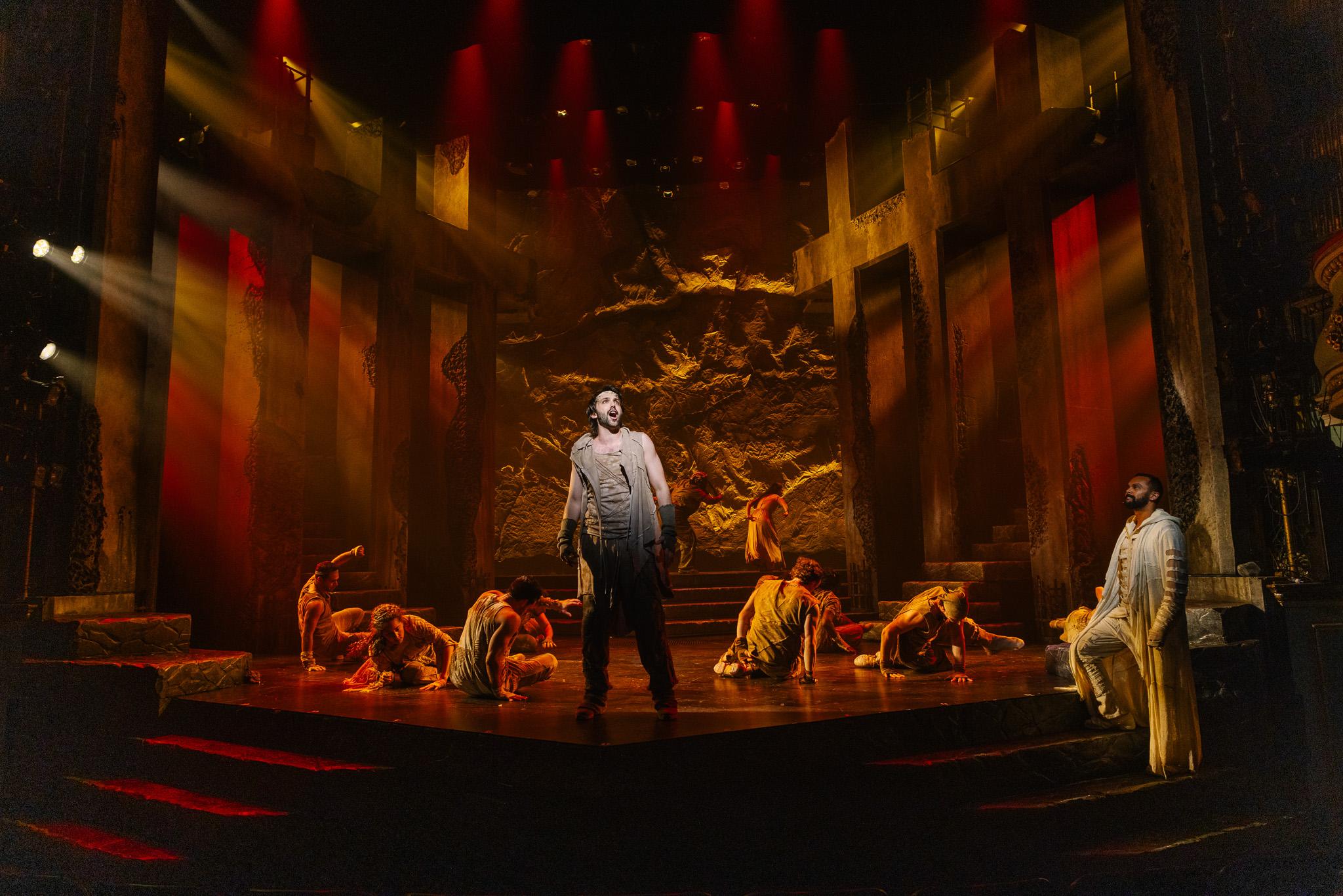 Shane Lonergan as Simon, standing centre stage in warm light with the company kneeling around him